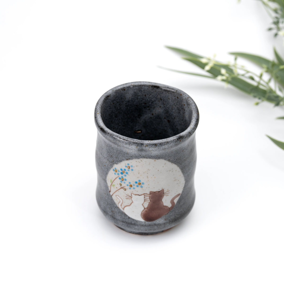 Blue-flower Kutani cat yunomi beside a leafy branch.