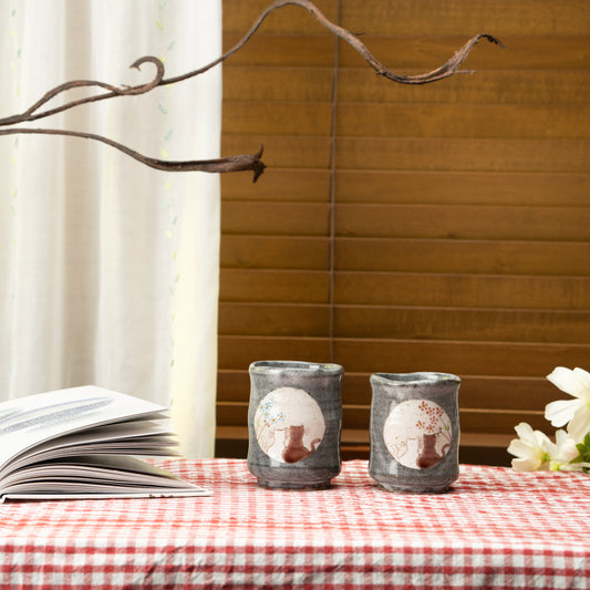 Kutani cat yunomi pair on a red checked tablecloth beside an open book and white flowers.