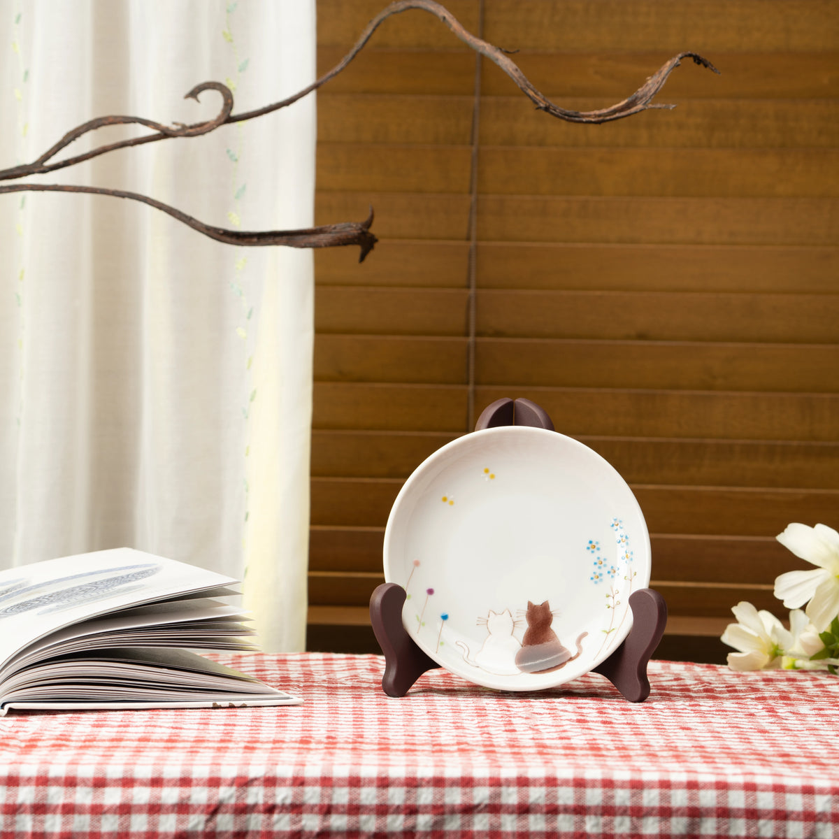Cat plate displayed on a stand on a red checked tablecloth beside a book.