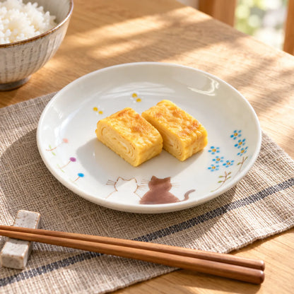 White Kutani Hidamari Cats plate holding two pieces of tamagoyaki, with the painted cats and blue flowers visible at the rim