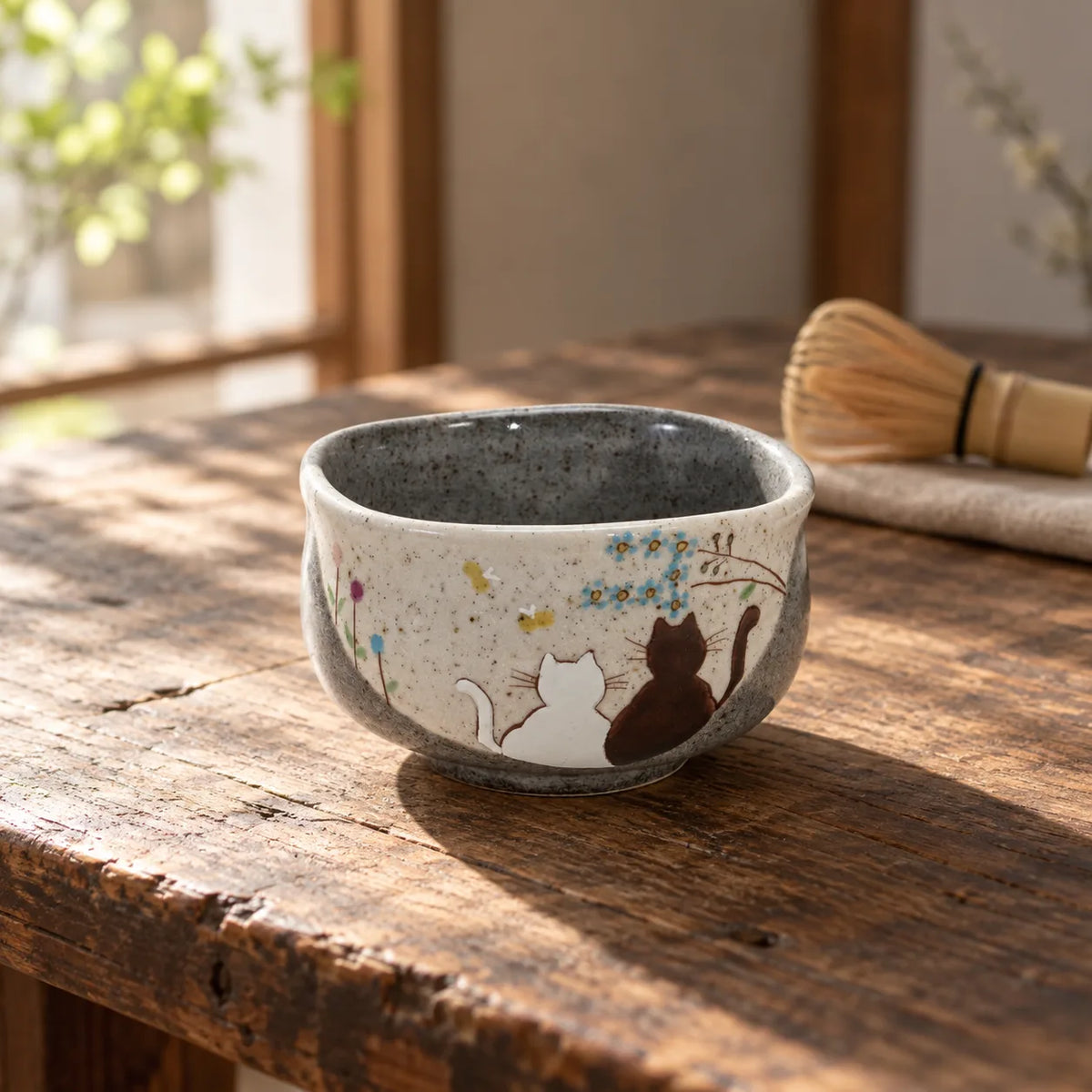 Iroe Yu Hidamari matcha bowl with two painted cats on a sunlit wooden table, with a bamboo whisk nearby.