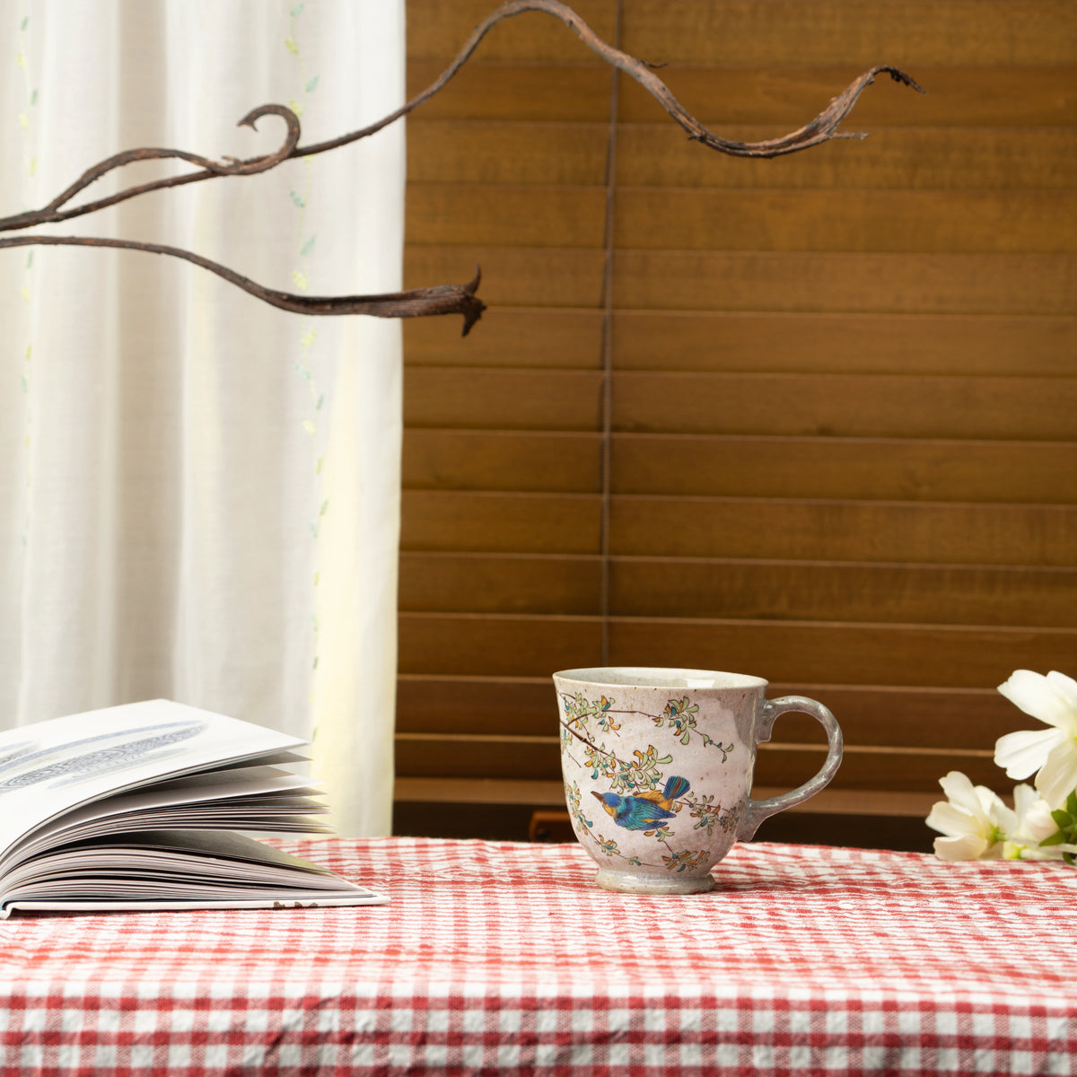 Kutani kingfisher mug on a red checked tablecloth beside an open book, white flowers and a wooden blind.