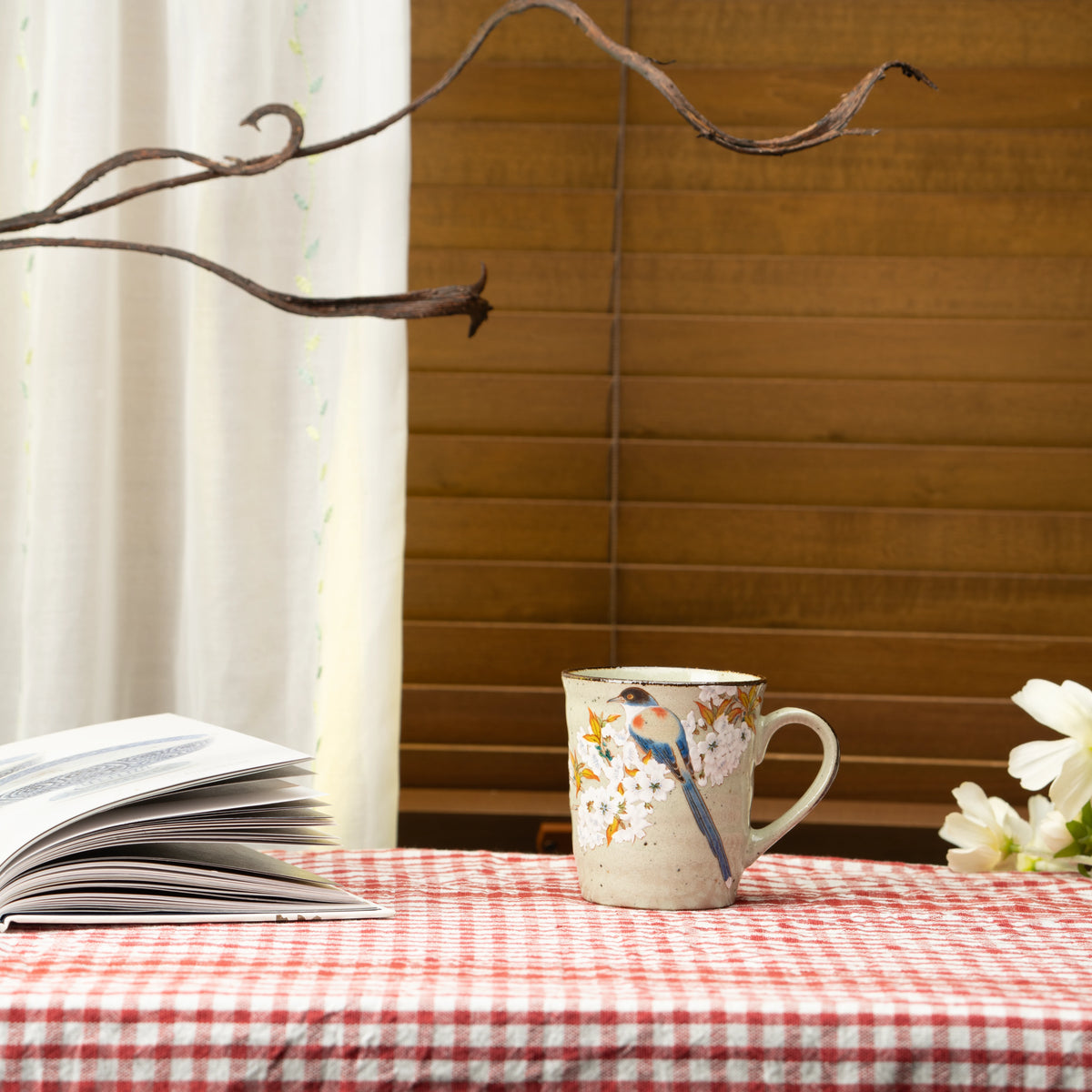 Bird and cherry blossom mug on a red checked tablecloth beside a book and flowers.