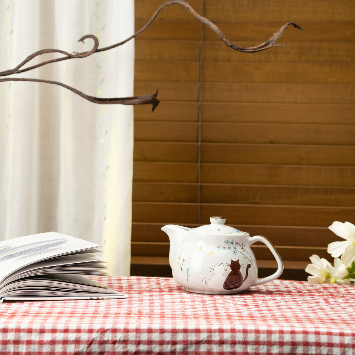 Kutani cat teapot on a red checked tablecloth beside a book and flowers.