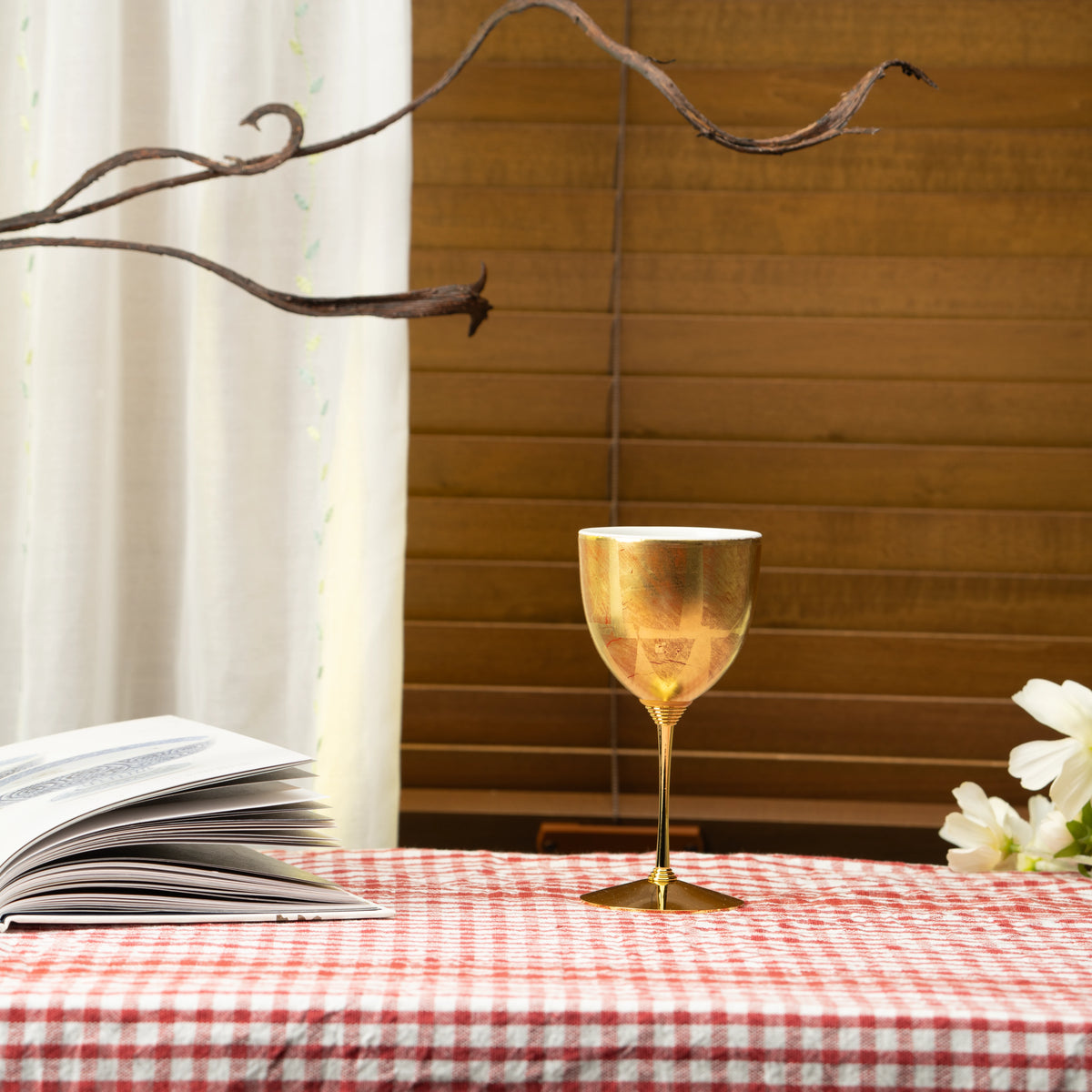 Gold wine cup on a red checked tablecloth beside a book and flowers.