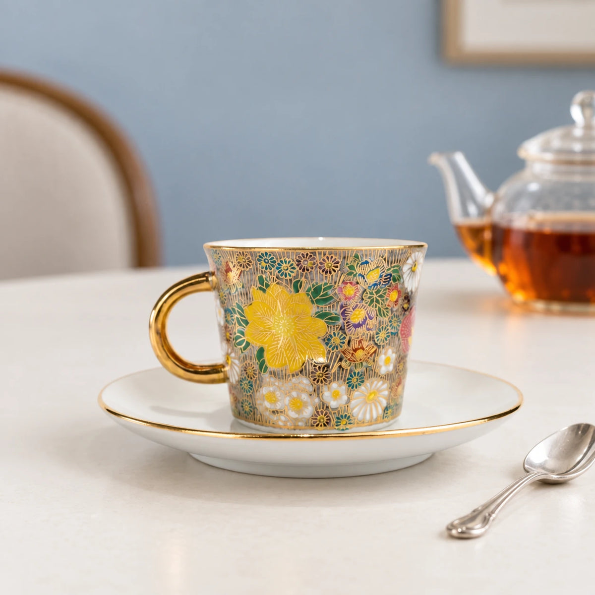 Kutani honkin hanazume floral cup and gold-rimmed saucer on a pale tea table, with a glass teapot in the background.