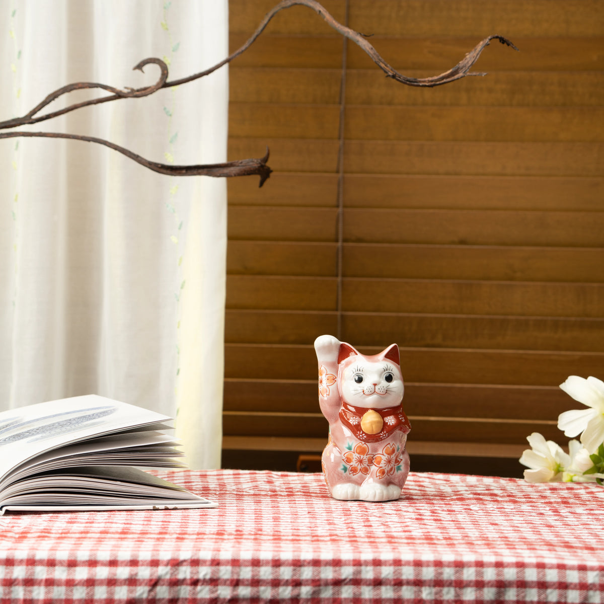 Pink cherry blossom lucky cat on a red checked tablecloth beside a book.
