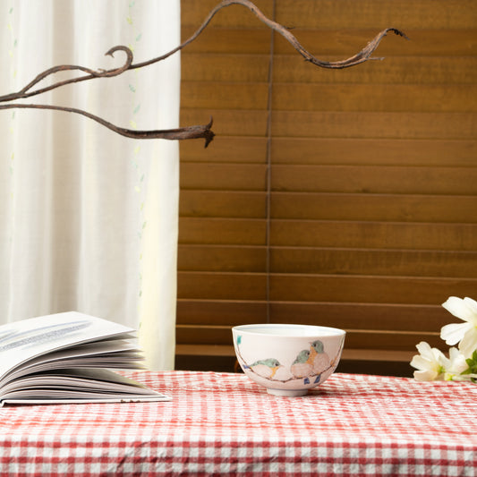 Bird rice bowl on a red checked tablecloth beside a book and flowers.