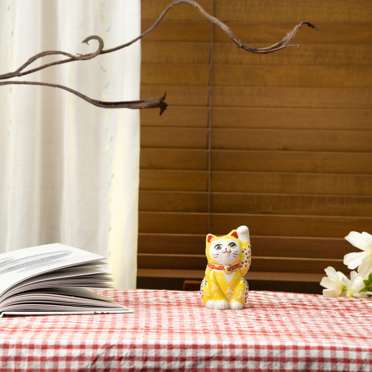 Yellow lucky cat on a red checked tablecloth beside a book.