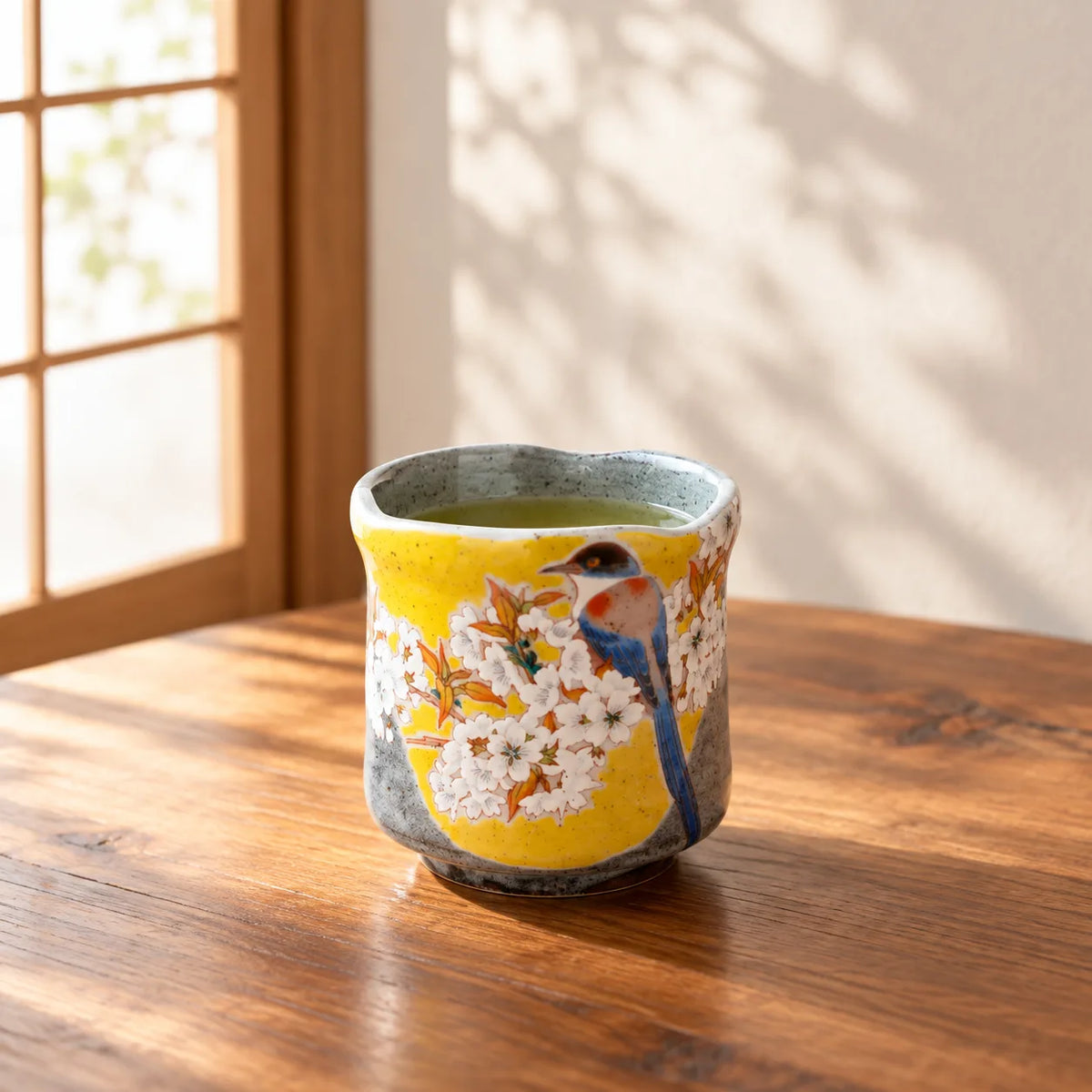 Kutani Yoshidaya yunomi with blue bird and white cherry blossoms holding green tea by a window