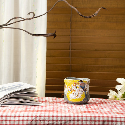 Kutani bird and cherry blossom yunomi on a red checked tablecloth beside a book and flowers.