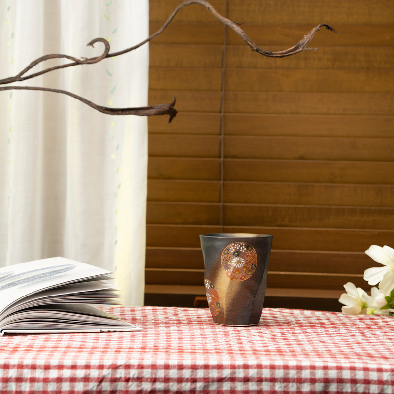 Black floral Kutani cup on a red checked tablecloth beside a book and white flowers.