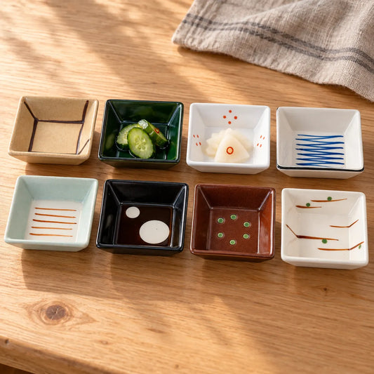 Eight small square Mino porcelain bowls with distinct patterns on a wooden table, two holding pickles and one holding sauce