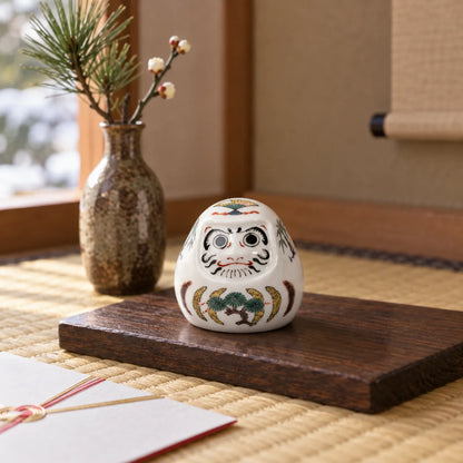 Miniature Kutani Sanyū Daruma with painted eyes, pine motif and a fan on its crown, displayed beside winter branches on tatami