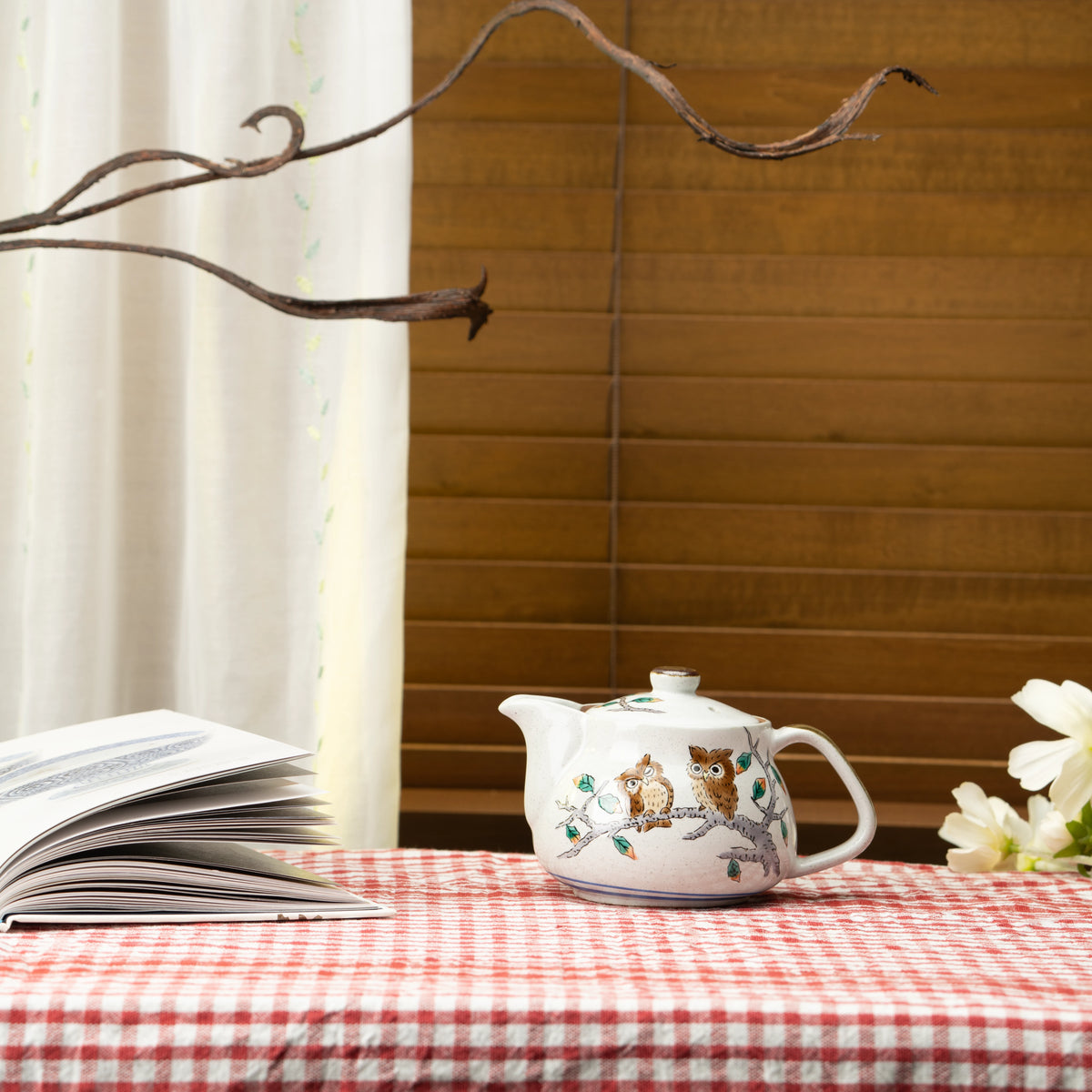 Kutani owl teapot on a red checked tablecloth beside an open book and white flowers.