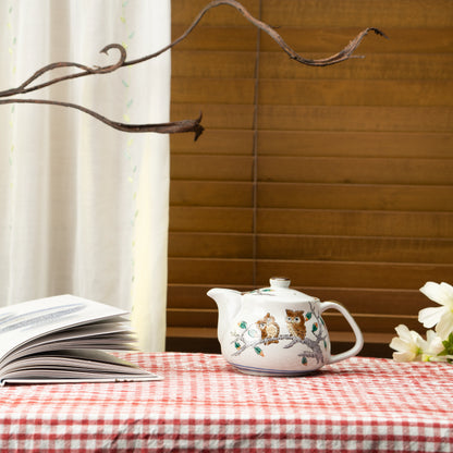 Kutani owl teapot on a red checked tablecloth beside an open book and white flowers.