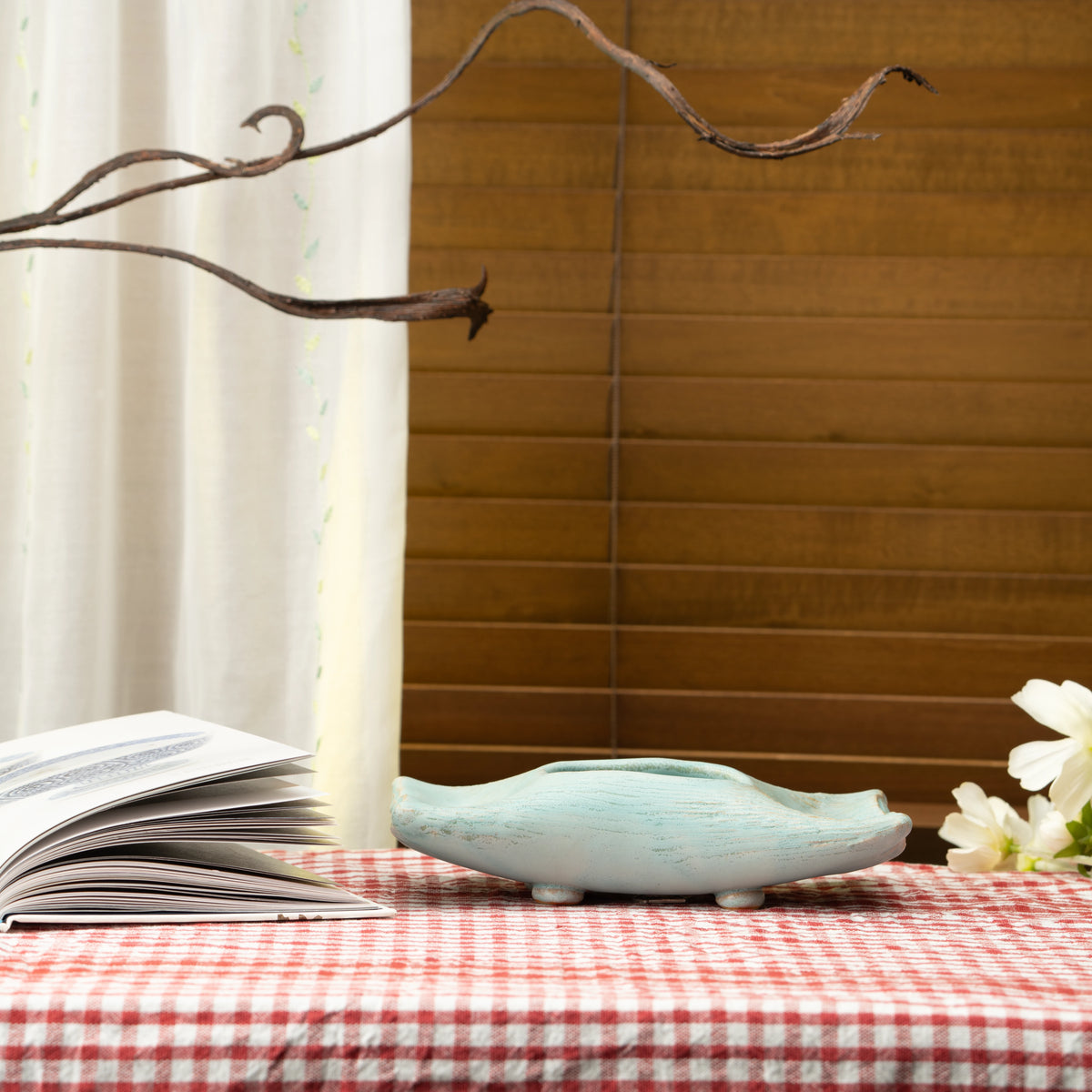 Pale blue Hechimon vase on a red checked tablecloth beside an open book and white flowers.