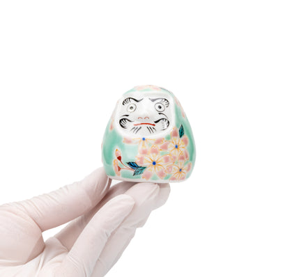 Small turquoise cherry blossom daruma held in a gloved hand.
