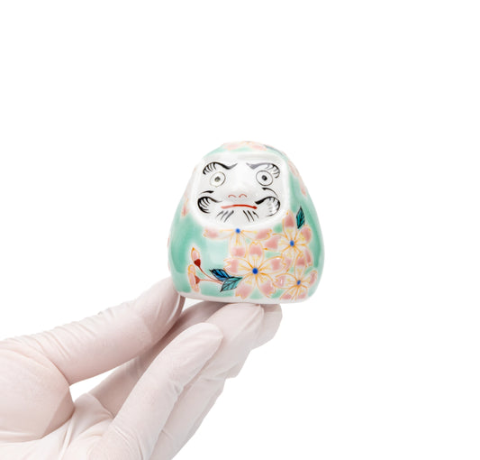 Small turquoise cherry blossom daruma held in a gloved hand.