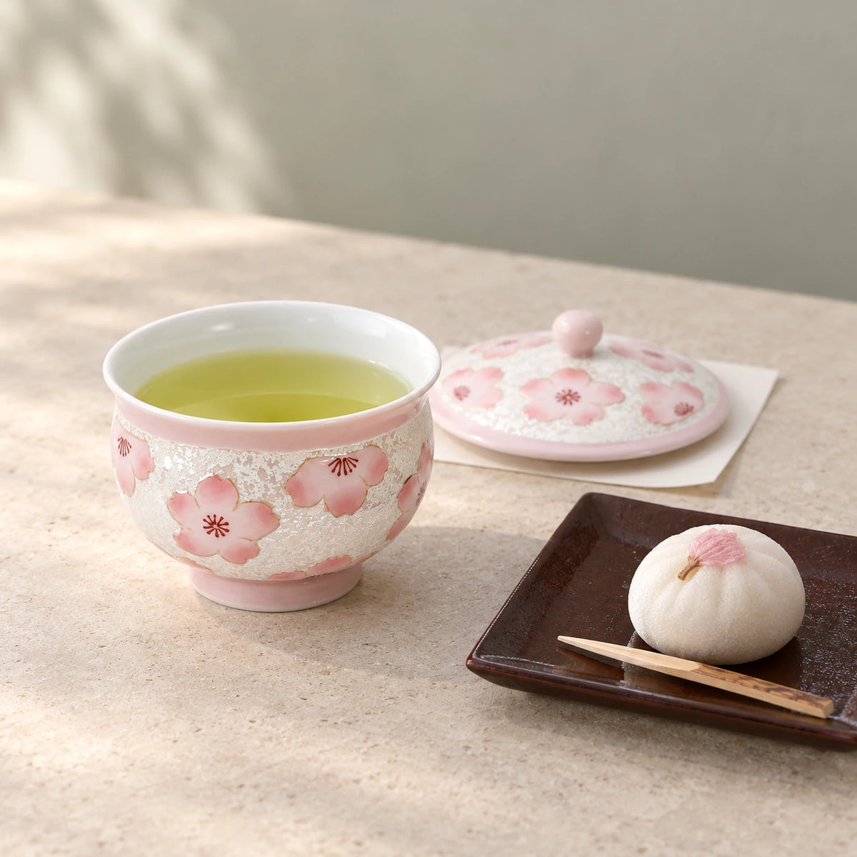 Pink cherry blossom teacup filled with green tea, its matching lid set beside it, and a small sweet on a dark plate.