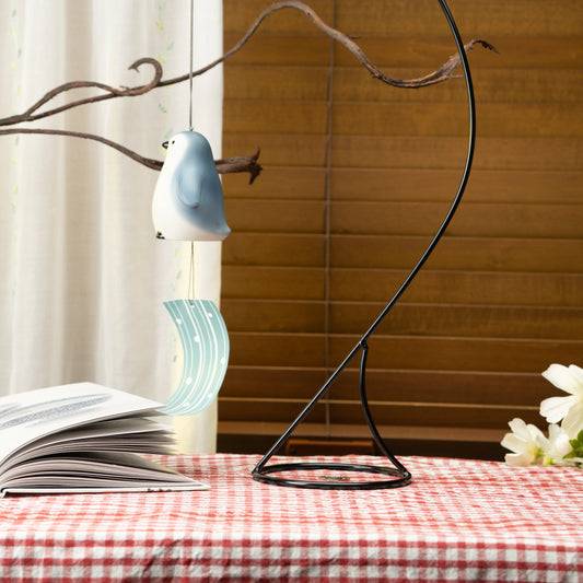 Penguin wind chime with pale blue paper strip hanging from a stand beside a window.