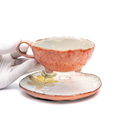 Orange floral cup held above its matching saucer in a gloved hand.