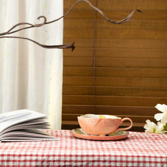 Orange cup and saucer on a red checked tablecloth beside a book and flowers.