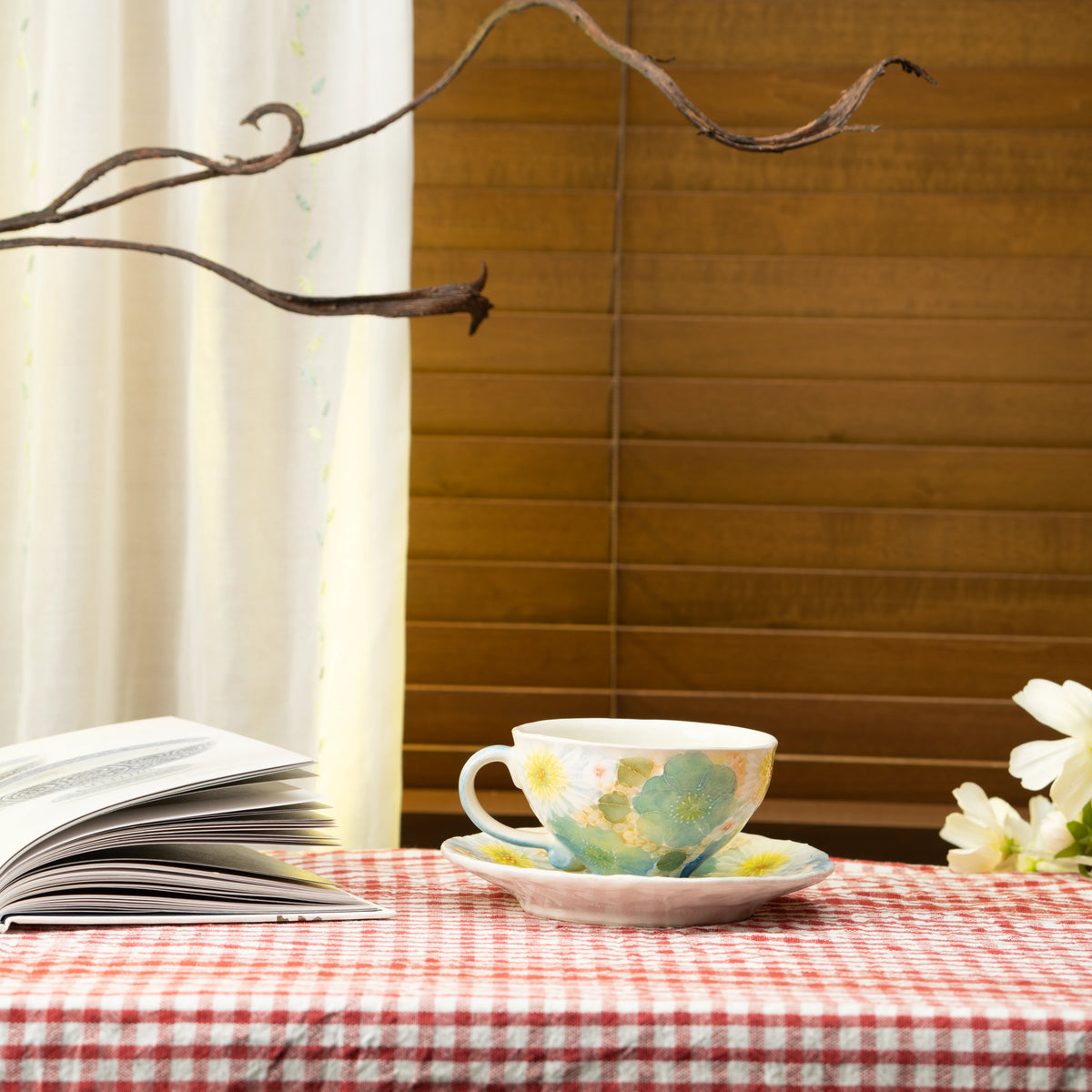 Blue floral cup and saucer on a red checked tablecloth beside a book.