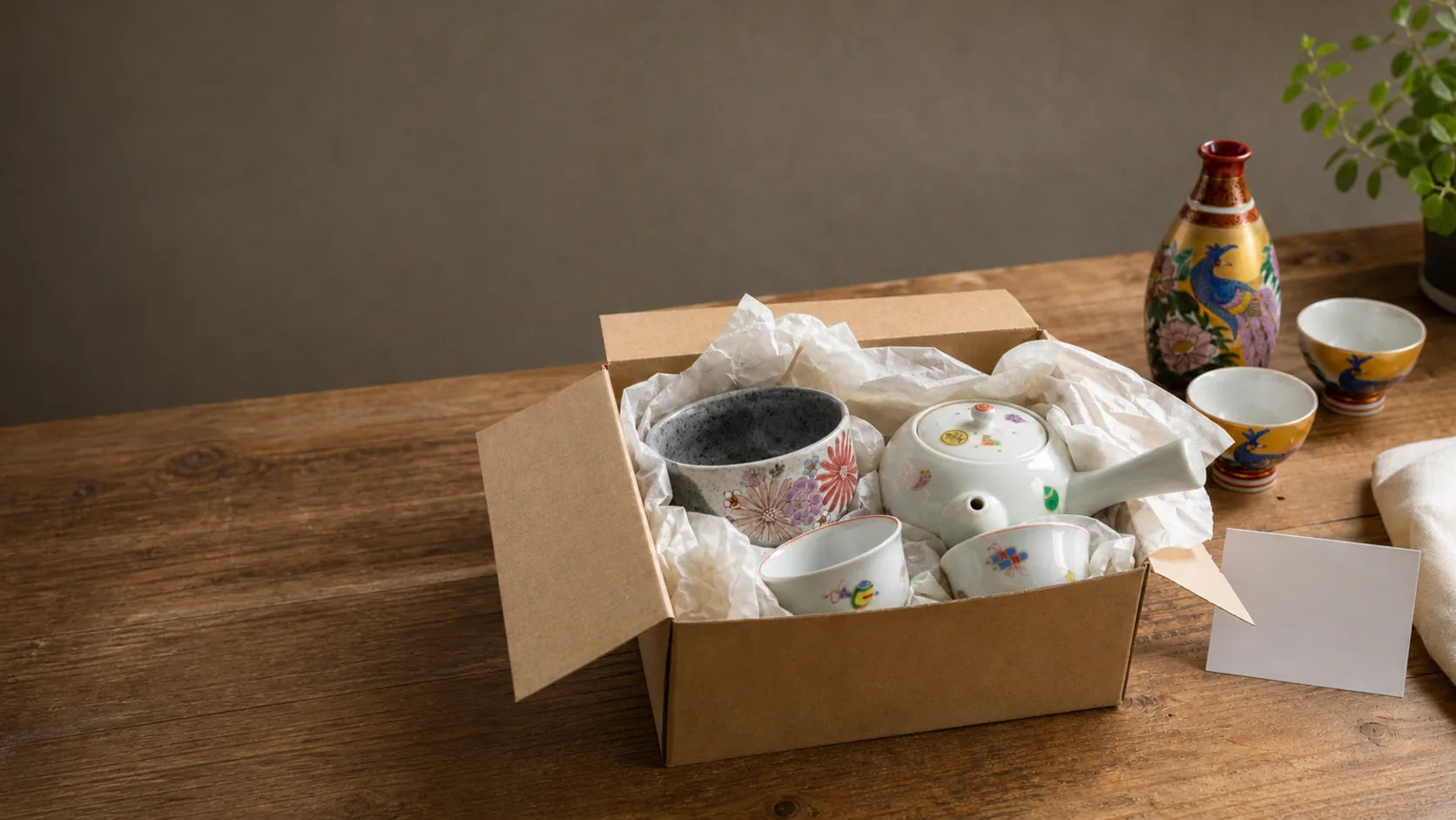 An open parcel with wrapped Japanese ceramics and a blank thank-you card on a warm wooden table.
